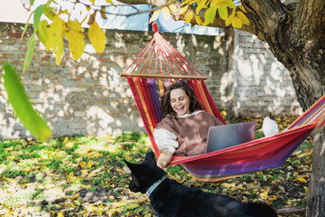 Young happy woman freelancer with laptop and dog in hammock at backyard of country house on autumn...