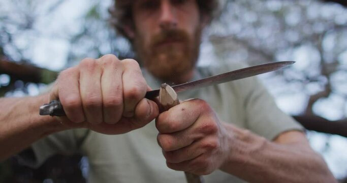 Caucasian male survivalist cutting point in stick to make fire drill at camp in wilderness