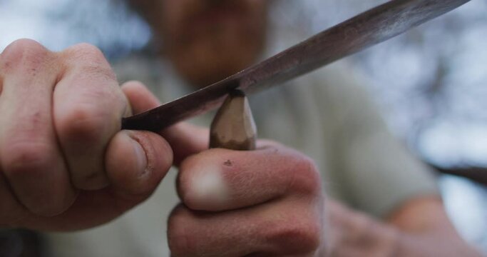 Caucasian male survivalist cutting point in stick to make fire drill at camp in wilderness