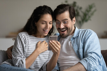 Head shot close up of happy couple looking at smartphone screen, reading unexpected good news in...