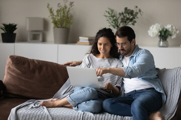 Happy couple using laptop, relaxing sitting on couch together, smiling beautiful woman and man in glasses looking at computer screen, chatting or shopping online, spending leisure time with device - obrazy, fototapety, plakaty