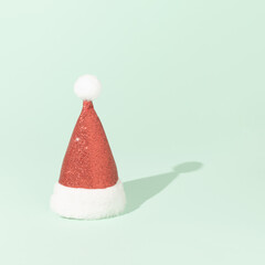 Red glitter Santa Claus hat with sunlight shadow against bright green background. Minimal Christmas concept. New Year party aesthetic. Winter holidays celebration idea.