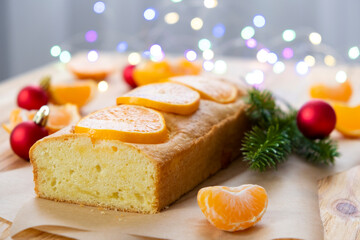 Loaf of tangerine cake with pieces of mandarin, fir branch, Christmas balls and lights on rustic wooden background. Tasty dessert for winter holidays. Merry Christmas and Happy New Year atmosphere.