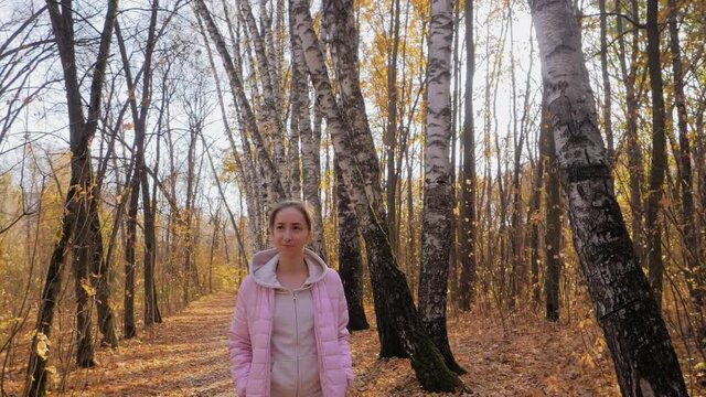 Slow motion: young woman walking in autumn park, forest - wide angle steadicam shot, front view. Sun shines through trees with sun lens flares. Active outdoor lifestyle, leisure time, freedom concept