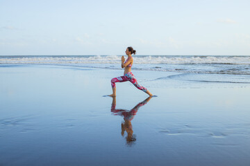 Beach yoga. Slim Caucasian woman practicing Virabhadrasana II, Warrior II Pose. Strong body. Healthy lifestyle. Water reflection. Yoga retreat. Outdoor exercise. Copy space. Bali