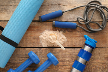 Measuring scoop with powder, fitness mat, skipping rope, bottle with protein shake and dumbbells on wooden background