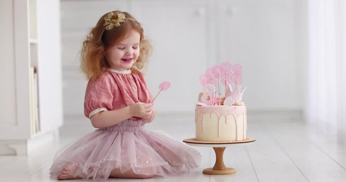 a three years old baby girl sitting next to her birthday cake, decorated with sugar lollipops, mastic crown and icing
