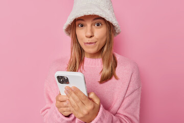 Cropped shot of pretty lovely young woman waits for call from boyfriend bites lips and has intriguing expression wears hat and casual jumper isolated over pink background watches video online