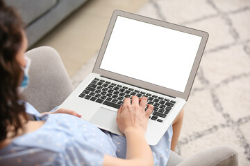 Young pregnant woman in medical mask using laptop at home