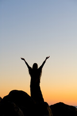 Woman standing on top of big rocks and raising her hands towards the sunset sky