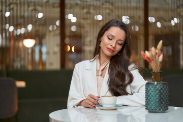 Elegant caucasian woman in white blazer sitting in cafe and drinking cappuchino coffee