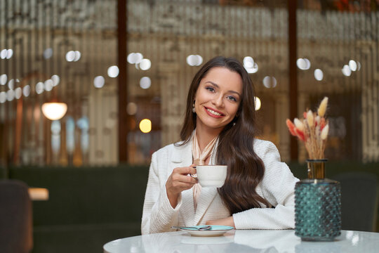 Elegant caucasian woman in white blazer sitting in cafe and drinking cappuchino coffee