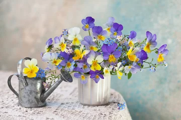 Fototapeten A bouquet of pansies and forget-me-not flowers in a vase and a watering can on a table with a tablecloth against the background of a colored wall. Romantic postcard, blur, selective focus. © tachinskamarina