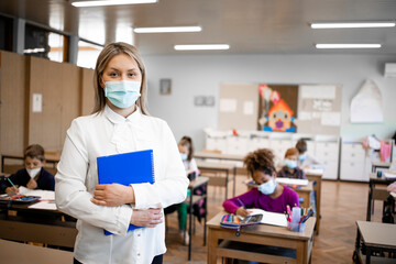 Portrait of school teacher during corona virus pandemic wearing face mask in classroom with children in background. - obrazy, fototapety, plakaty