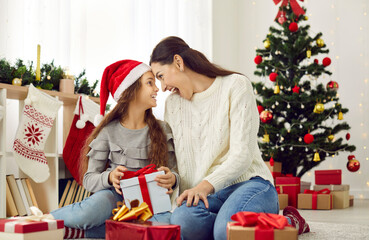Obraz na płótnie Canvas Merry Christmas. Young cheerful mother and her daughter enjoy Christmas and New Year holidays at home. Portrait of funny family having fun sitting among gifts in living room with Christmas decor.