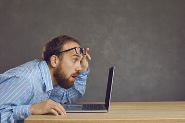 Side view of a man with a surprised expression sitting at a table and staring at a laptop screen....