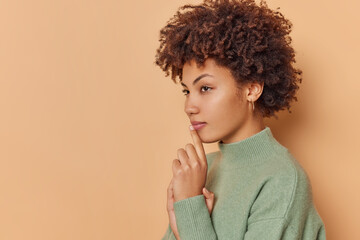 Profile shot of thoughtful curly haired woman thinks about something with mysterious intrigued expression wears casual jumper isolated over beige background blank copy space for your advert.