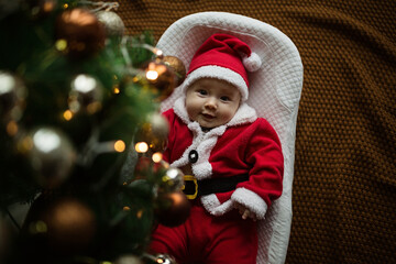 Closeup portrait of newborn baby. Cute Caucasian baby girl 4-5 months old in Santa costume lying on knitted cozy blanket in cocoon near decorated fir tree. Merry Christmas xmas and happy new year 2022