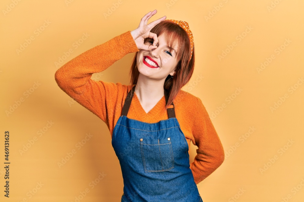 Wall mural Redhead young woman wearing apron smiling happy doing ok sign with hand on eye looking through fingers