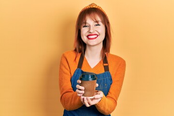 Redhead young woman wearing waitress apron holding take away cup of coffee smiling with a happy and cool smile on face. showing teeth.