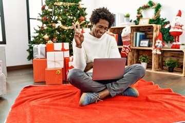 Young african american man using laptop sitting by christmas tree smiling with happy face winking...