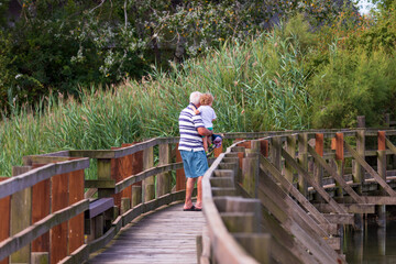 Nonno con nipote in braccio passeggia su un piccolo ponte nella riserva naturale nella laguna del mare adriatico a marano lagunare. Passeggiata estiva tra la natura. - obrazy, fototapety, plakaty