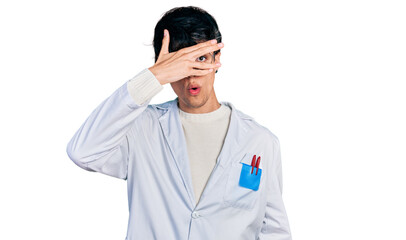 Handsome hipster young man wearing doctor uniform peeking in shock covering face and eyes with hand, looking through fingers afraid