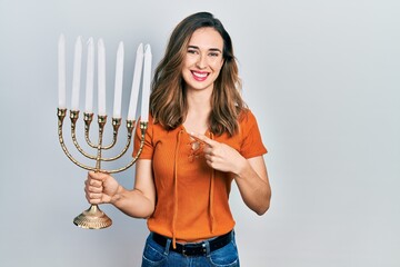 Young hispanic girl holding menorah hanukkah jewish candle smiling happy pointing with hand and finger