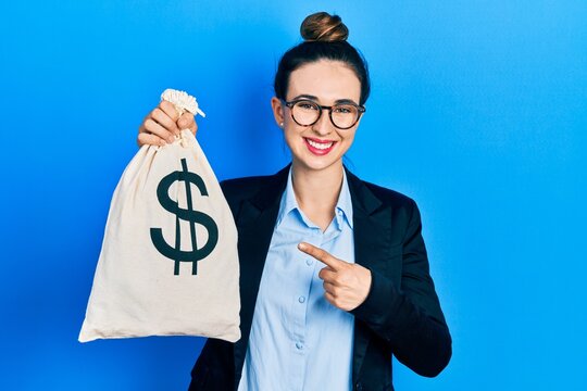 Young hispanic girl wearing business clothes holding dollars bag smiling happy pointing with hand and finger