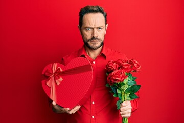 Middle age man holding anniversary present and bouquet of flowers skeptic and nervous, frowning...