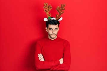 Young hispanic man wearing cute christmas reindeer horns skeptic and nervous, disapproving expression on face with crossed arms. negative person.