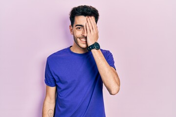 Young hispanic man wearing casual t shirt covering one eye with hand, confident smile on face and surprise emotion.