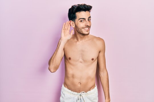 Young hispanic man wearing swimwear shirtless smiling with hand over ear listening an hearing to rumor or gossip. deafness concept.