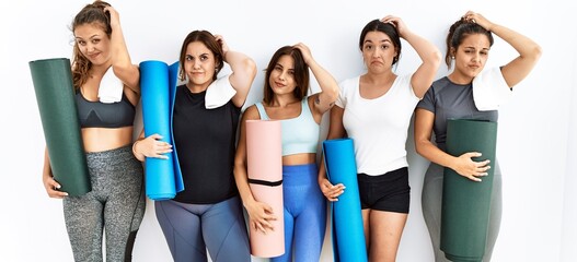 Group of women holding yoga mat standing over isolated background confuse and wondering about question. uncertain with doubt, thinking with hand on head. pensive concept.