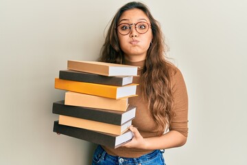 Young hispanic girl holding a pile of books puffing cheeks with funny face. mouth inflated with air, catching air.