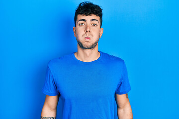 Fototapeta premium Young hispanic man wearing casual blue t shirt puffing cheeks with funny face. mouth inflated with air, crazy expression.