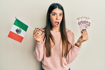 Young hispanic woman holding mexico flag and mexican pesos banknotes afraid and shocked with...