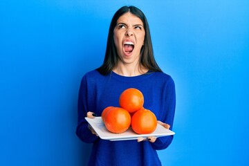 Young hispanic woman holding plate with fresh oranges angry and mad screaming frustrated and furious, shouting with anger looking up.