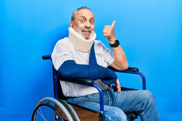 Handsome senior man with beard sitting on wheelchair with neck collar an arm sling pointing thumb...