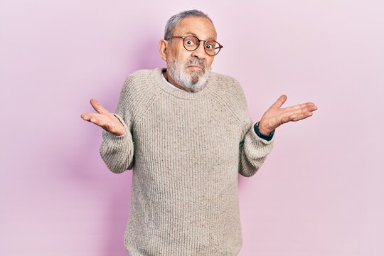 Handsome senior man with beard wearing casual sweater and glasses clueless and confused expression with arms and hands raised. doubt concept.