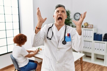 Middle age doctor man at the clinic with a patient crazy and mad shouting and yelling with aggressive expression and arms raised. frustration concept.