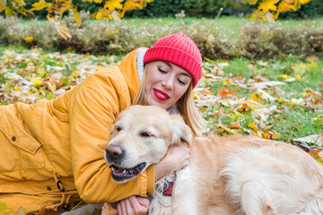 woman in warm clothing hugs her retriever lying on the grass among  leaves autumn.