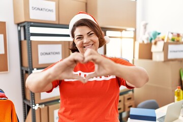 Middle age hispanic woman wearing volunteer t shirt and christmas hat smiling in love showing heart symbol and shape with hands. romantic concept.