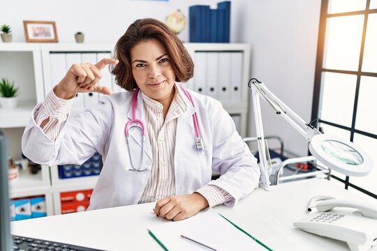 Middle age hispanic woman wearing doctor uniform and stethoscope at the clinic smiling and confident gesturing with hand doing small size sign with fingers looking and the camera. measure concept.