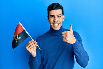 Handsome hispanic man holding angola flag smiling happy and positive, thumb up doing excellent and approval sign