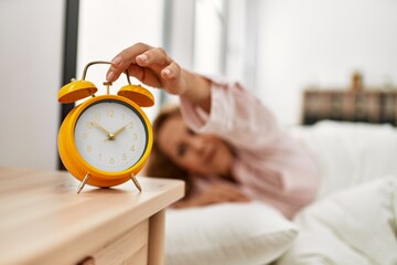 Middle age caucasian woman turning off alarm clock lying on the bed at bedroom.