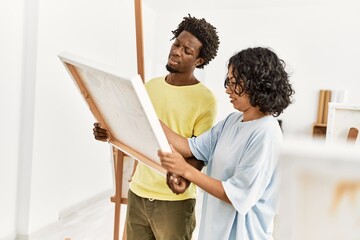African american artist couple with serious expression holding canvas at art studio.