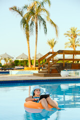 A Jewish woman in a sun hat in the pool in a swimming circle with a cocktail and a laptop on a background of palm trees.
