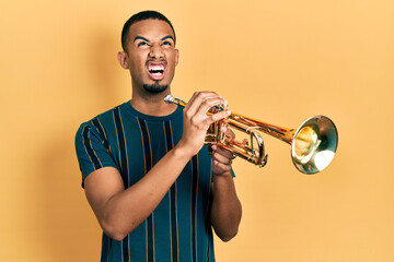 Young african american man playing trumpet angry and mad screaming frustrated and furious, shouting...