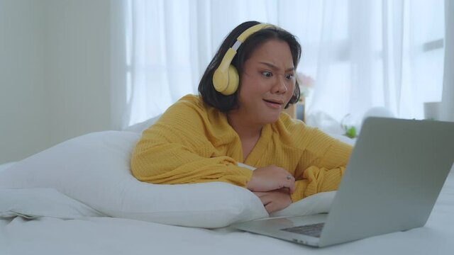 A fat woman in a yellow shirt and headphones lies in bed watching a movie in her bedroom. She shows shock or surprise while watching the movie. Holiday activities lifestyle concept.